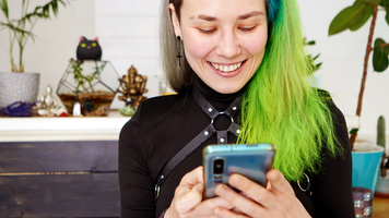 Image of young punk girl no makeup with bizarre hairstyle smiling while using smartphone in indie interior